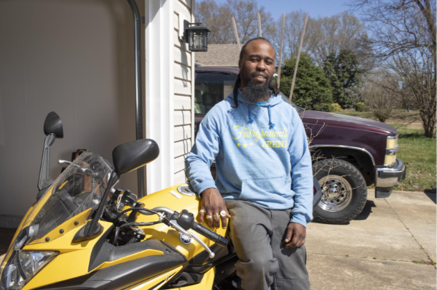 A photo of Demetre, a Self-Help member,
        leaning on a motorcycle in the driveway of his home 