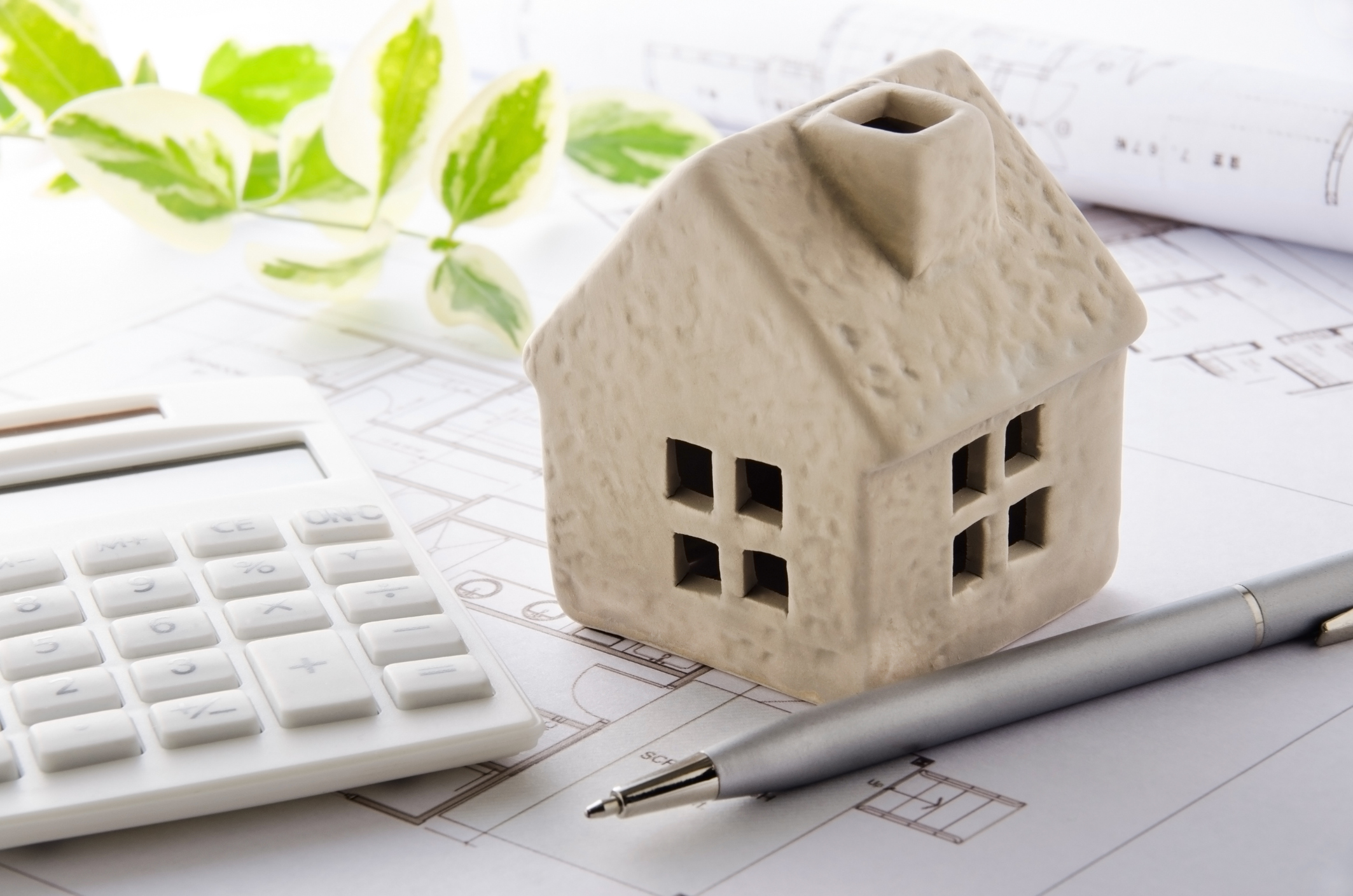 small cardboard model of a house on top of blueprints with a calculator to its left and a green vine behind them and a silver pen in front of the house