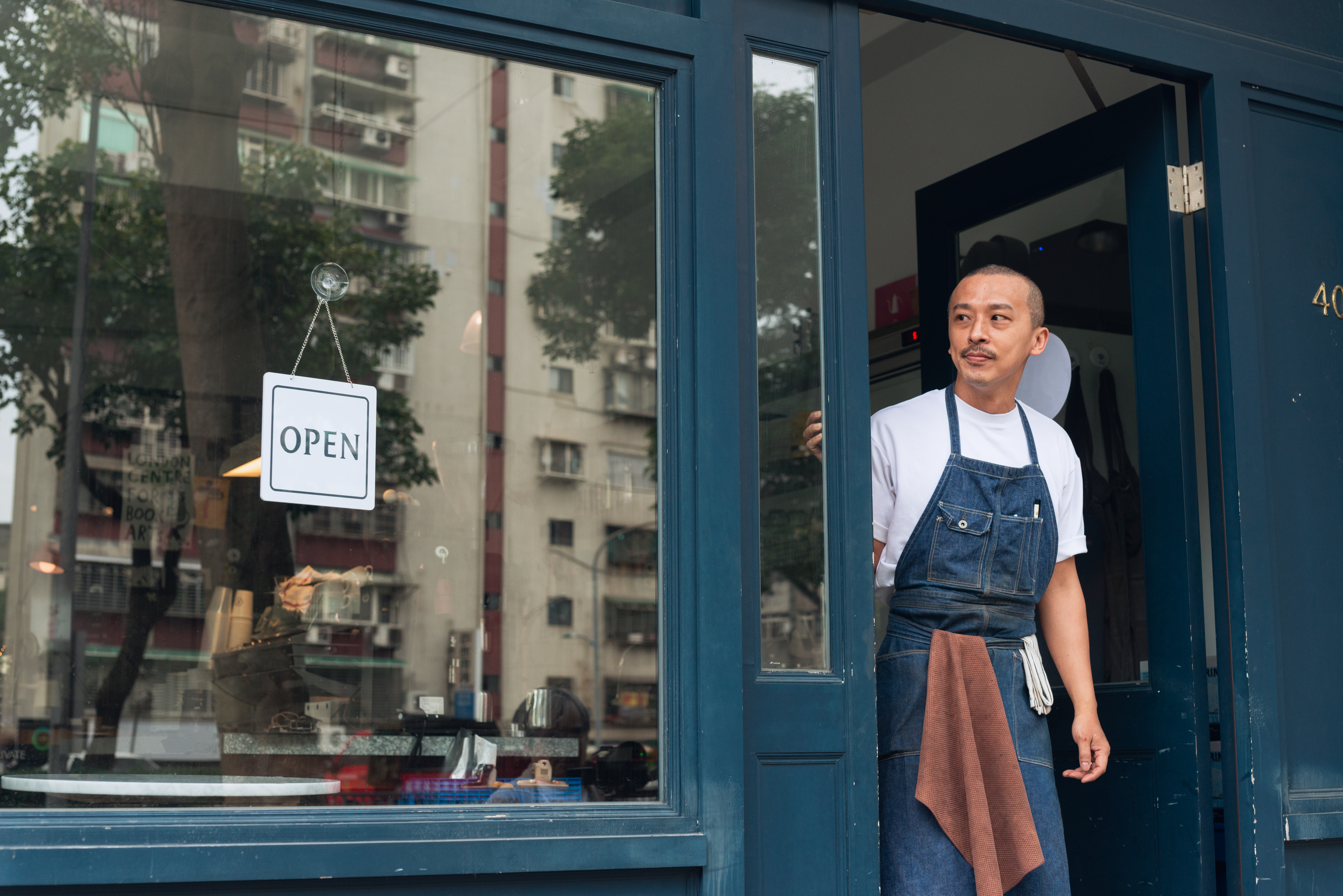 Asian man wearing an apron standing in the doorway of a business that has a sign that reads