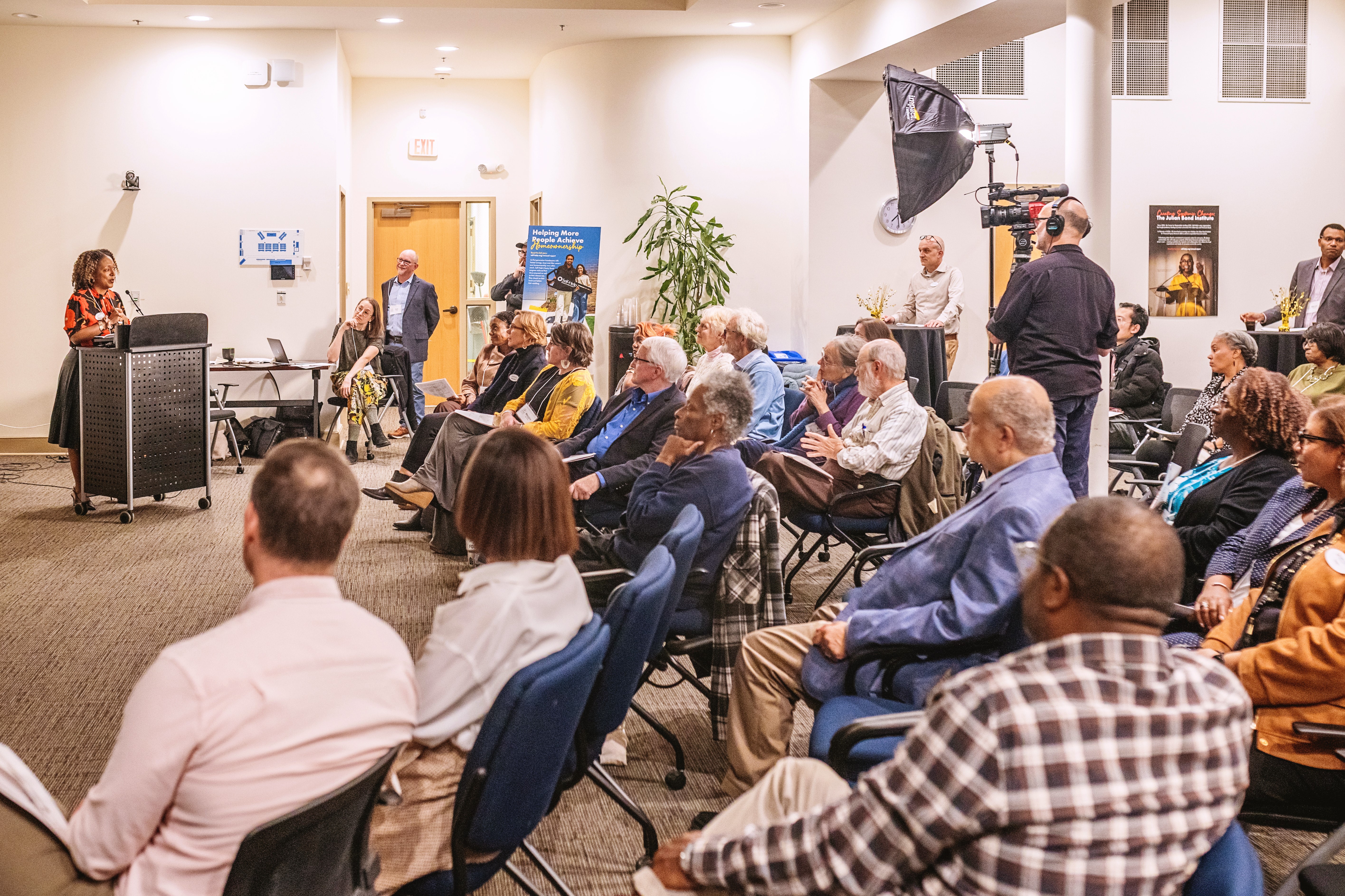A photograph of a group of people sitting in chairs viewing and listening to a person speak at a podium in the front of the room